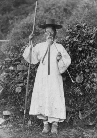 A Korean man in traditional robe and boots (hanbok), wearing a horse hair and bamboo hat (gat), holding a staff and smoking a pipe. 1885.jpg, sept. 2019