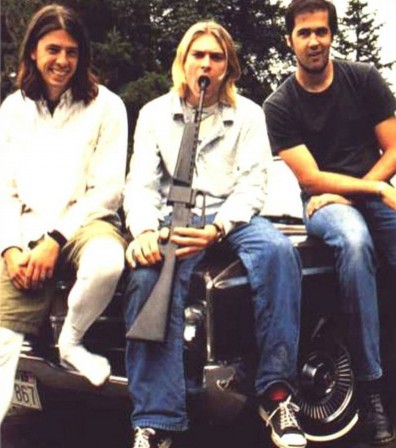Kurt Cobain pose avec son groupe assis sur une voiture, avec un fusil dirigé vers sa bouche 1992.jpg, juil. 2021