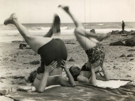 sur la plage, deux femmes font la chandelle, au bord de l'eau, un homme regarde l'horizon