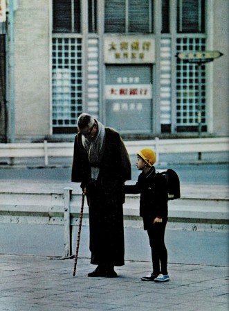 un écolier japonais portant un cartable donne la main à un vieil homme en tenue traditionnelle muni d'une canne