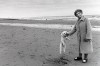 sur une plage déserte, une vieille femme en manteau d'hiver, une laisse à la main,  pose en train de faire sauter son caniche
