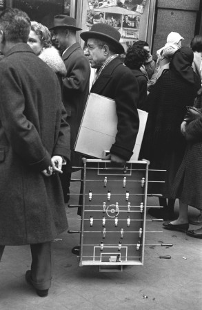 dans une rue animée, un homme mur porte un chapeau et un jeu de babyfoot