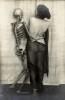 un squelette amoureux enlace la taille d'une femme de dos, uniquement vêtue d'un voile transparent