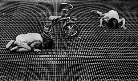 à côté d'un tricycle abandonné, deux enfants accroupis collent leur visage à la grande grille d'aération recouvrant le sol pour voir au travers