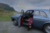 sur un chemin de bord de mer, un ancien modèle de Volvo bleue arrêtée, le corps de la conductrice brune pendant par la porte ouverte, la tête près du gravier, les yeux ouverts