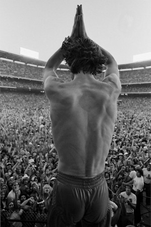 Mick Jagger, de dos, les mains jointes en forme de tour Eiffel au dessus de sa tête, devant la foule assemblée dans un stade