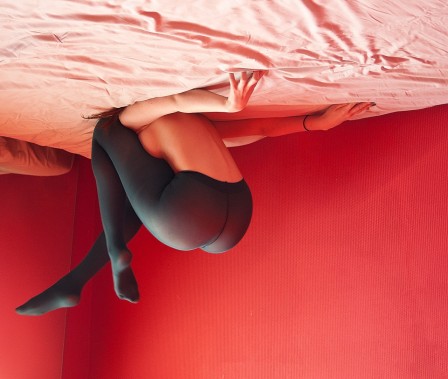 dans une chambre rouge, sur son lit au plafond, une femme en collant noir exerce sa souplesse